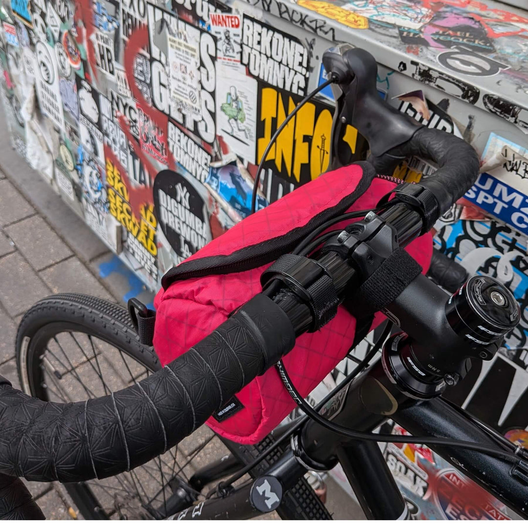 Close-up of a hot pink ecopak handlebar bag against a wall covered in stickers.